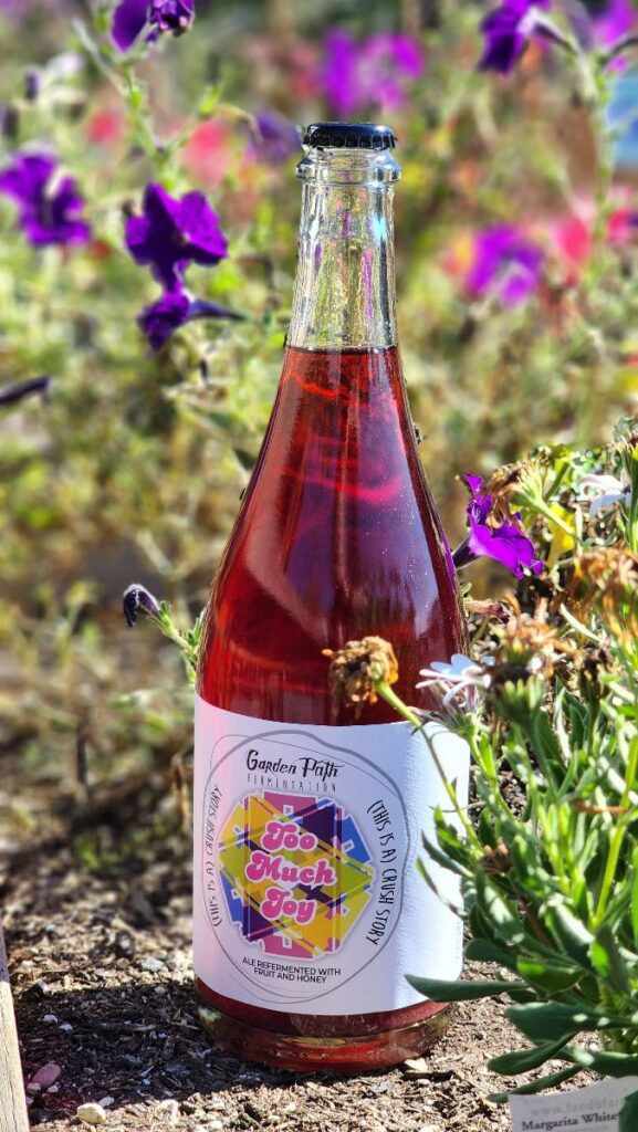 A bottle of (This is) A Crush Story made by Garden Path fermentation. It is sitting among some flowers in the sun.